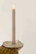 Load image into Gallery viewer, Chandelle allumée en cire d’abeille gaufrée dans un bougeoir en céramique claire, tenu dans une main sur fond crème.
